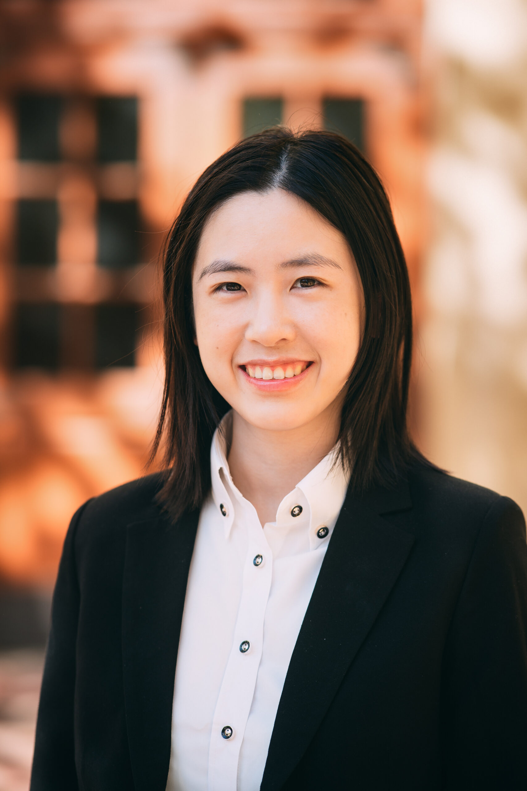Professor Chen smiling against a blurred light brown background wearing a black coat and white shirt